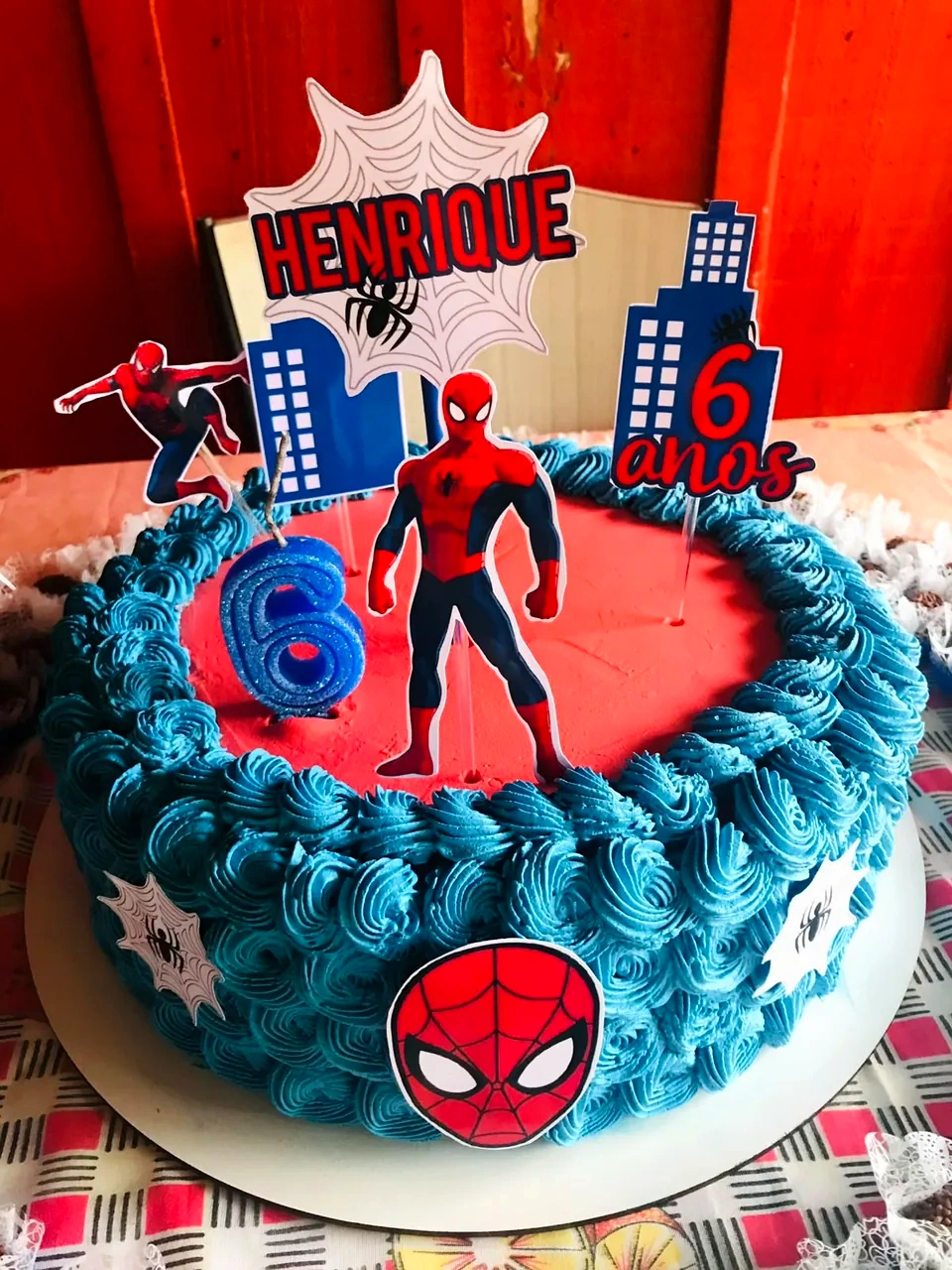 Bolo azul e vermelho decorado com toppers do Homem-Aranha, prédios, teia e vela em formato do número 6.