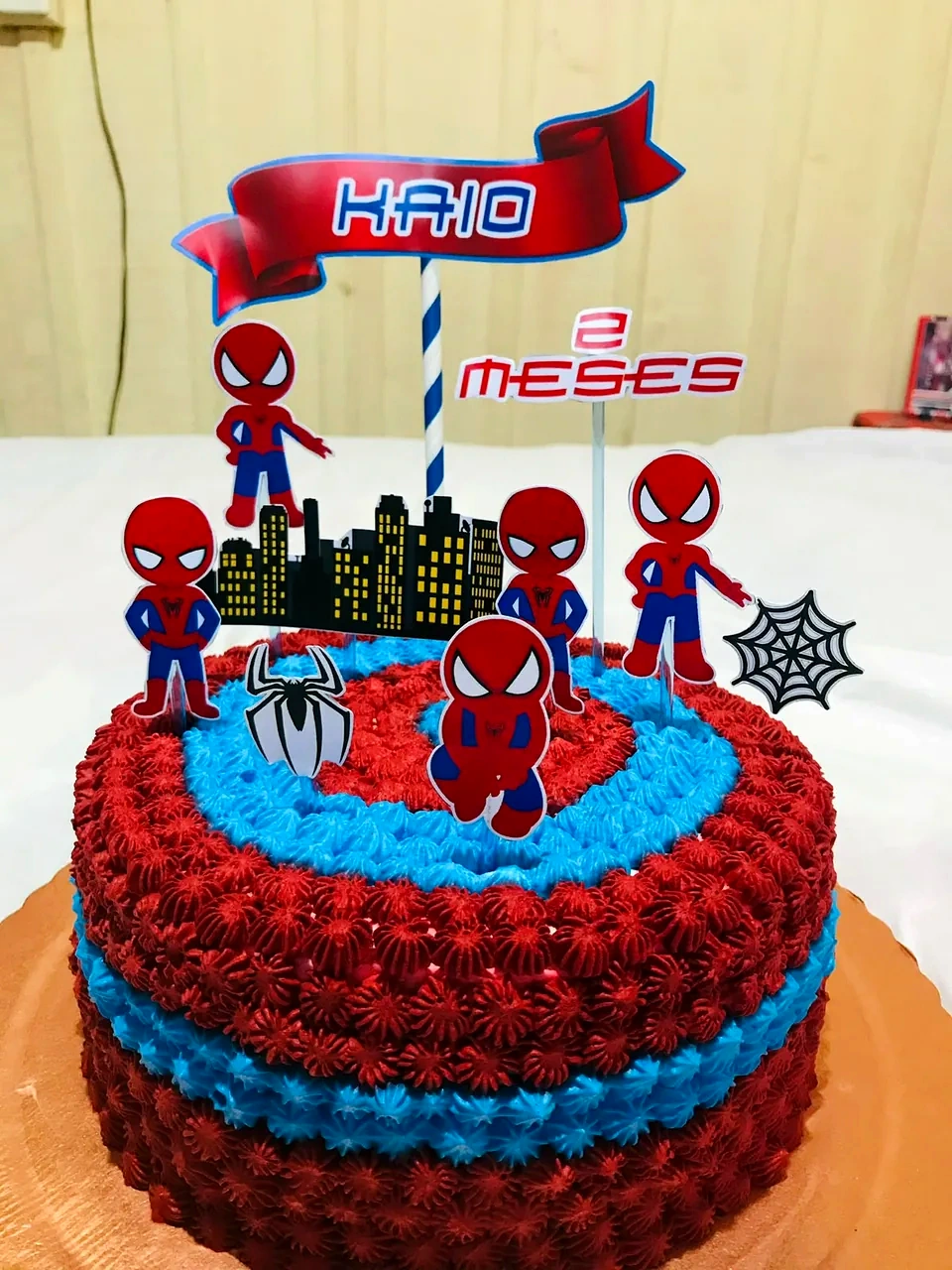 Bolo em camadas de bicos vermelhos e azuis, com toppers de pequenos Homem-Aranha, cidade e faixa com nome.