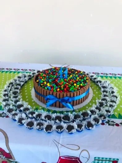 Bolo com laterais de barras de chocolate, laço azul e confeitos coloridos, cercado por docinhos em forminhas.