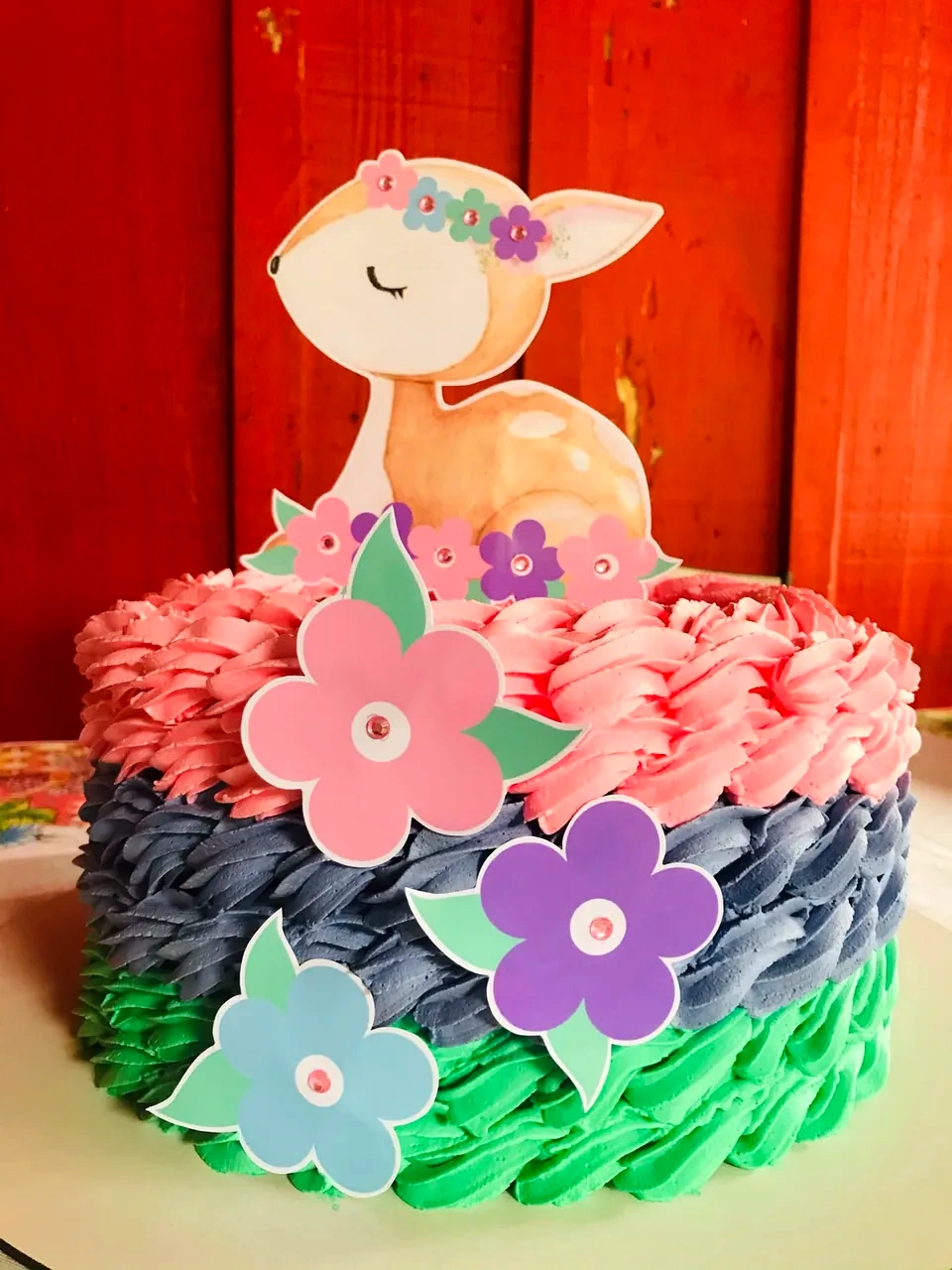 Bolo com três faixas de cobertura nas cores rosa, lilás e verde, decorado com flores coloridas e um topper de cervinho.