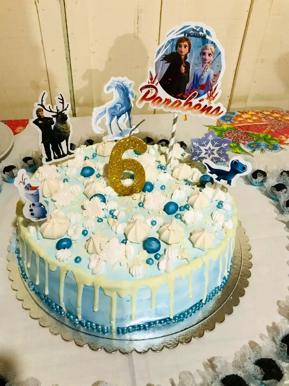 Bolo azul claro com escorrido branco, pérolas azuis, vela dourada com o número 6 e toppers de personagens de Frozen.