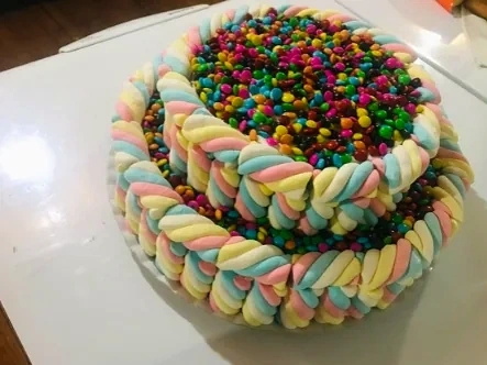 Bolo de dois andares com laterais de marshmallow trançado e topo coberto de confeitos coloridos.