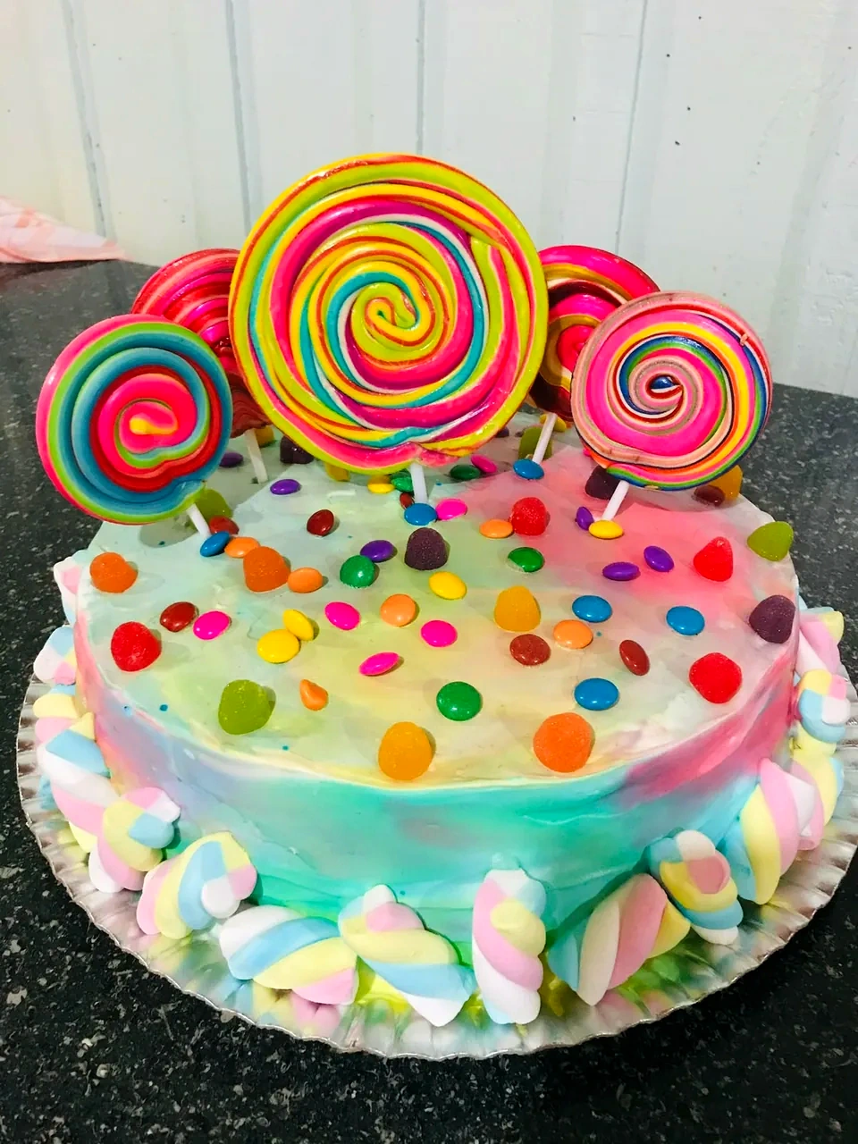 Bolo em tons pastel coberto de confeitos coloridos, com pirulitos gigantes no topo e marshmallows trançados na base.