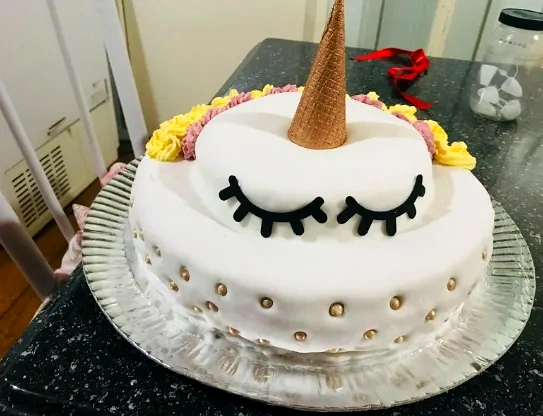 Bolo branco com detalhes dourados, cílios pretos, chifre dourado e flores amarelas, no tema unicórnio.