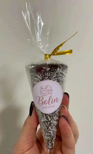Mão segurando um cone embalado em celofane com laço dourado e etiqueta Bolin Doces, com cobertura de chocolate e coco.