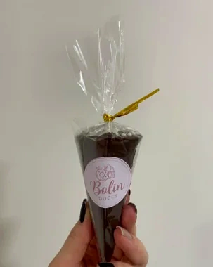 Mão segurando um cone de chocolate embalado em celofane, com laço dourado e etiqueta Bolin Doces.