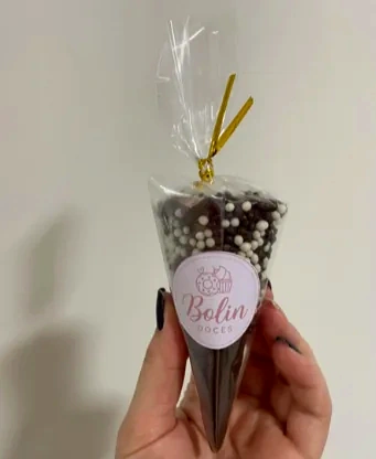 Mão segurando um cone embalado com laço dourado e etiqueta Bolin Doces, com cobertura de chocolate e bolinhas brancas.