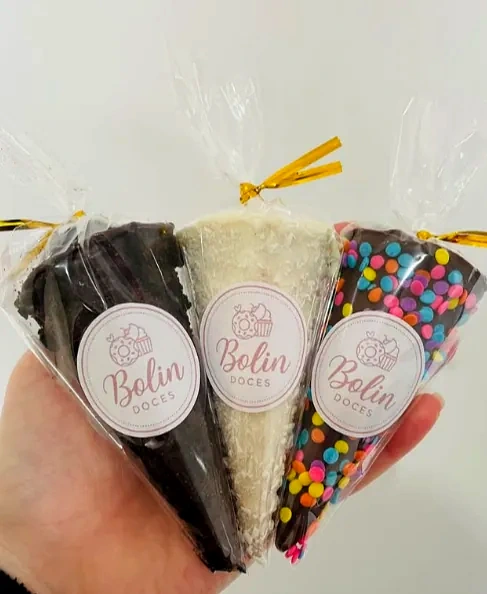 Mão segurando três cones embalados com etiqueta Bolin Doces: um de chocolate escuro, um branco com coco e um com confeitos coloridos.