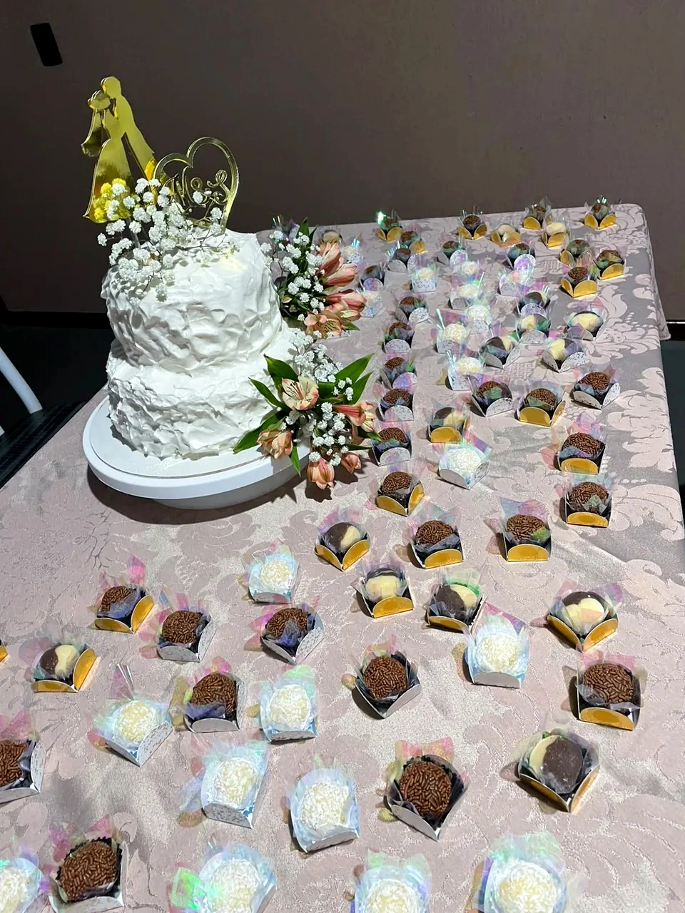 Mesa de festa com bolo de dois andares com flores e topper dourado, e docinhos em forminhas organizados à frente.
