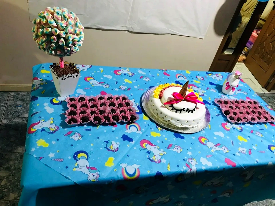 Mesa de festa com toalha azul de unicórnios, bolo decorado, docinhos em forminhas rosa e uma árvore de marshmallows.